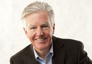 Smiling man with swept back grey hair, parted on the side, wearing open collar dress shirt, no tie and a dark jacket