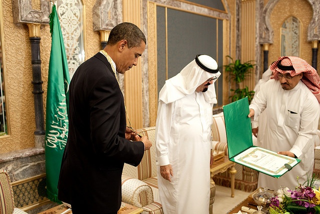 President Barack Obama in ornate room with Saudi flag and two men dressed in traditional Saudi garb