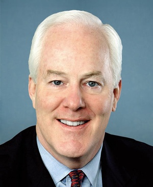 Head and shoulders shot of Sen. John Cornyn, white haired man with blue eyes wearing dark jacket, pale blue shirt and red print tie