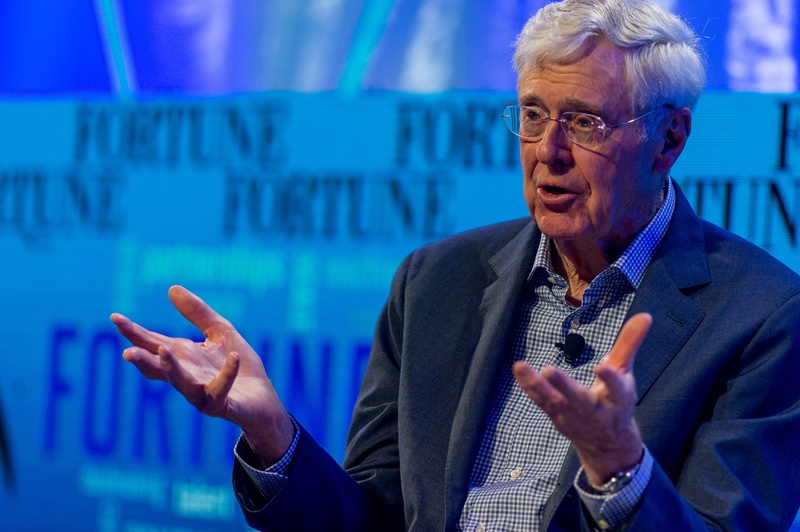 Charles Koch sitting on stage speaking with hands raised.