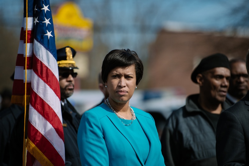DC Mayor Muriel Bowser standing next to an American flag with people in the background.