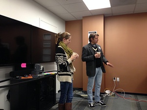 A man and woman dressed casually and holding coffee cups standing in front of a large black TV screen and speaking to an unseen audience 