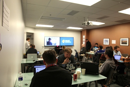 Photo of participants at PDF hackathon hunched over computers at tables in a conference room. Two large screens in the front show an image of a person and the blue and yellow logo of the Sunlight Foundation