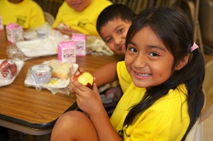 Kids eating school lunch, one smiling with a half-eaten apple
