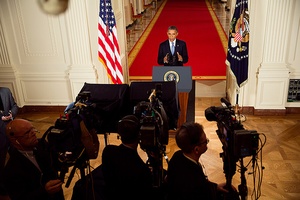 Image of President Obama standing before red carpet at press conference, with teleprompter and camera crew silhouetted in foreground