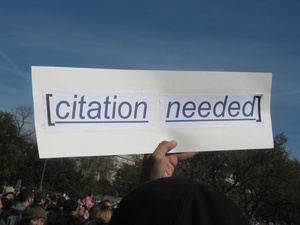 Hand holding a sign that reads "Citation Needed" within square brackets