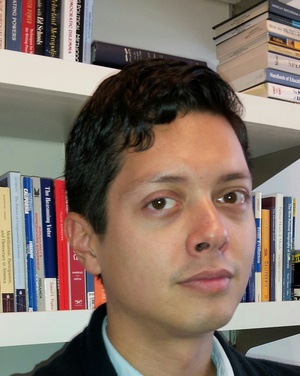 Head and shoulders shot of dark-haired young man standing in front of bookcases.