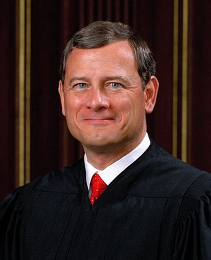 Head and shoulders shot of Chief Justice John Roberts, a brown haired, blue-eyed man smiling into the camera and wearing a black judicial robe with a white shirt collar and red tie visible underneath.