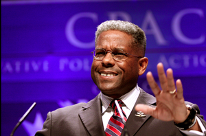 Picture of smiling, bespectacled African American man, waving in front of CPAC banner, in suit and red and blue striped tie