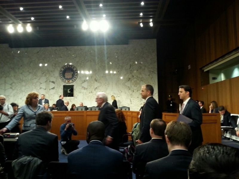 White haired man, followed by two men in suits, walking across Senate hearing room