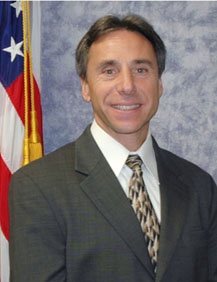 Waist up shot of smiling brown-haired man in brown suit with a U.S. flag behind him