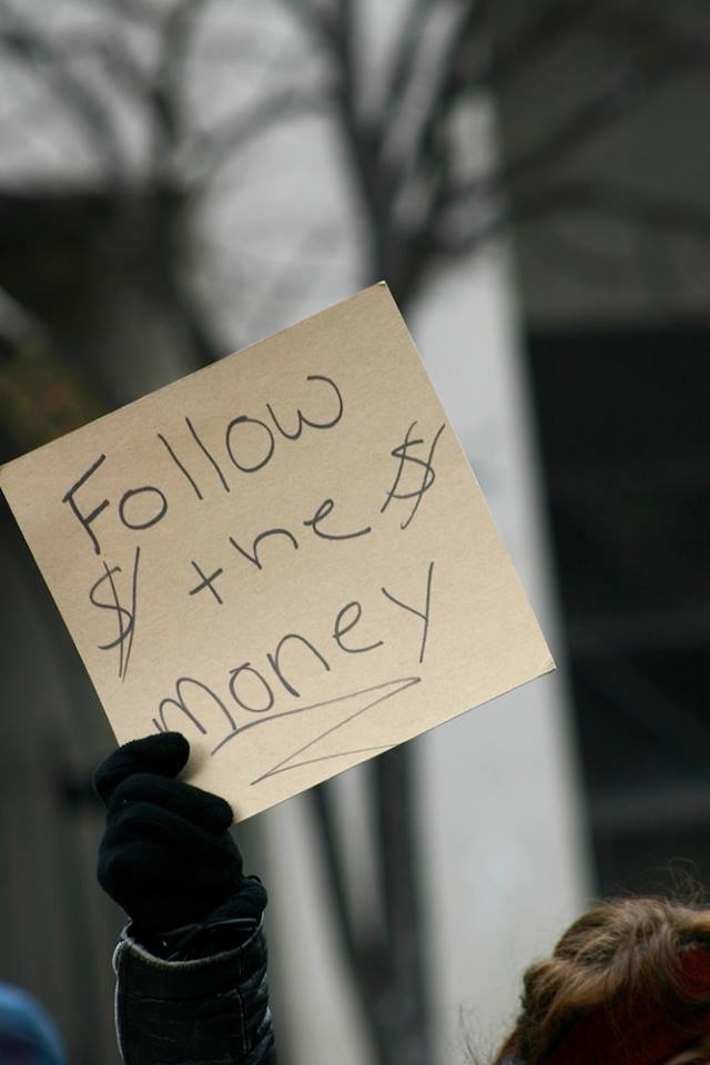 Someone at a demonstration holding up a sign that says follow the money.