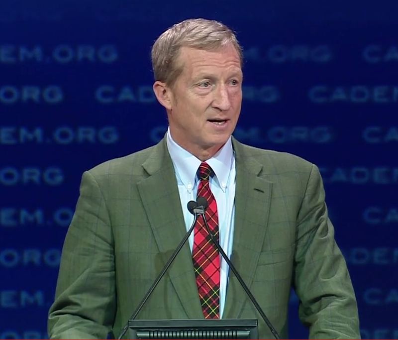 Tom steyer in green suit speaking in front of blue background.
