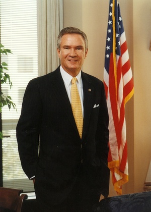 three-quarters portrait of John Breaux, wearing a dark suit in his former Senate office with an American flag to his right.