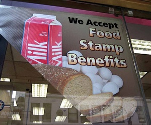 Sign in store window stating "we accept food stamp benefits" over a picture of eggs, a loaf of sliced bread and a carton of milk