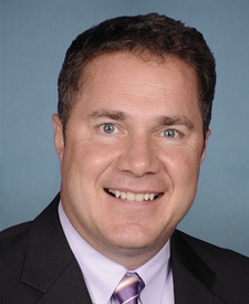 Head and shoulders shot of smiling dark-haired white man with blue eyes, wearing a lavender shirt, purple striped tie and black suitcoat 