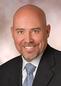 Head and shoulder shot of Tom MacArthur, bald man with salt and pepper goatee wearing dark suit jacket, white shirt and blue and yellow print tie