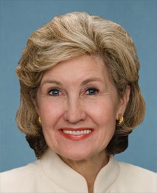 Head and shoulders shot of Kay Bailey Hutchison, white female with blonde hair wearing a white dress, gold earrings and lipstick